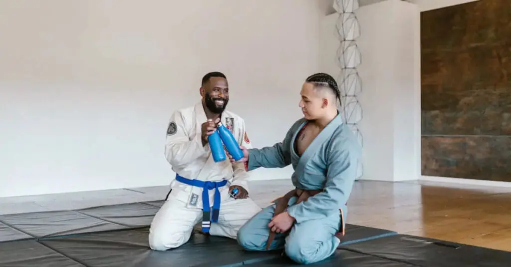 Two Brazilian jiu jitsu for beginners students in gis sitting on mats, smiling and clinking water bottles